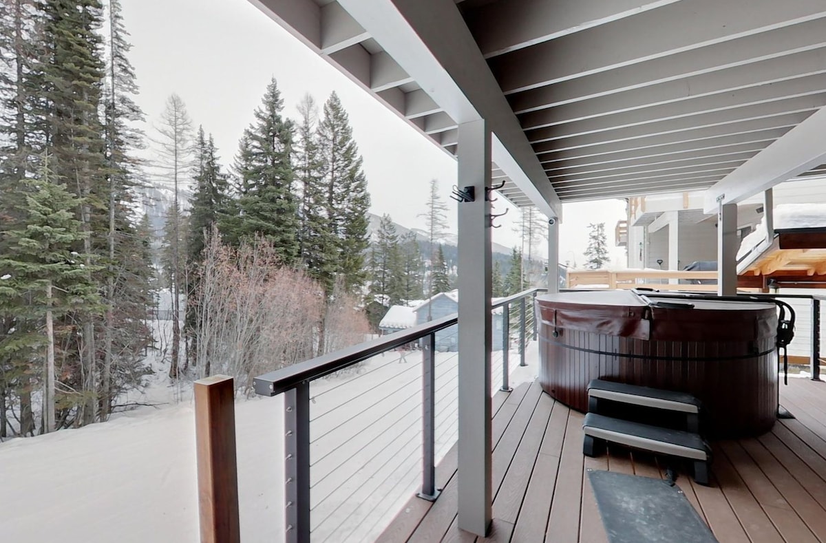 A hot tub is positioned on a wooden deck, surrounded by snow-covered trees and a tranquil winter landscape. The view showcases a serene atmosphere, with the snow blanketing the ground and mountain peaks in the distance.