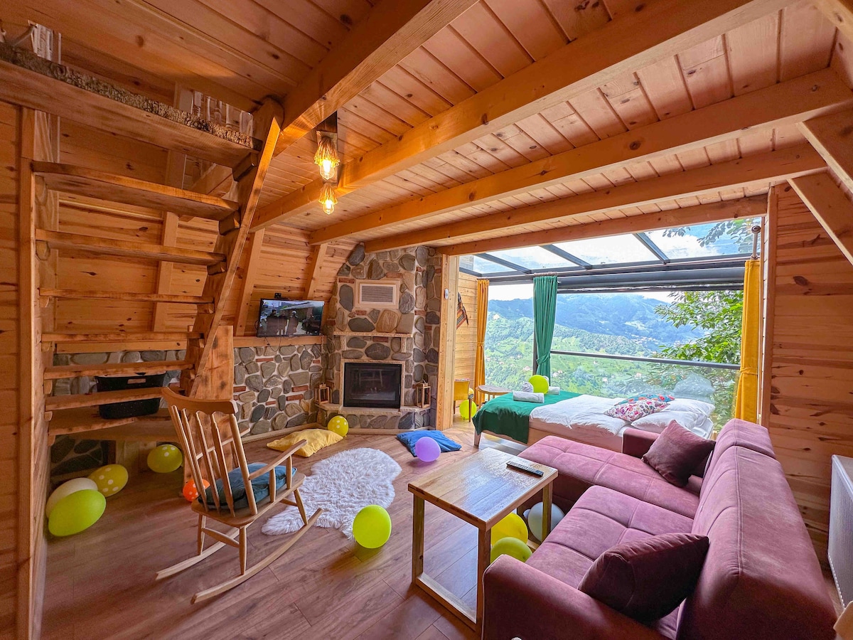 A wooden interior showcases a cozy living space with a soft rug and comfortable seating arrangements. A stone fireplace is centered in the room, while large glass doors provide views of the surrounding valley. Colorful balloons add a festive touch to the atmosphere.