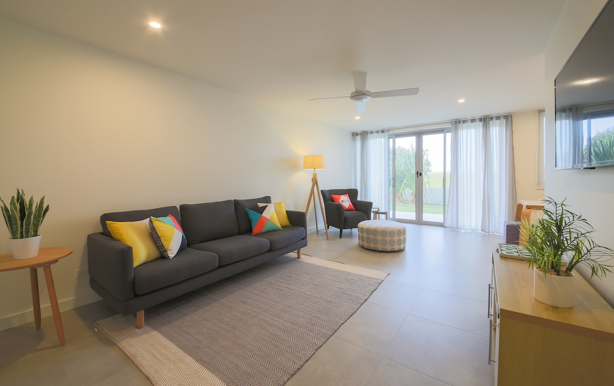 A spacious living area features a dark sofa adorned with colorful cushions, complemented by a round ottoman. Natural light streams through large sliding glass doors, which open to a view of the garden. A ceiling fan and warm lighting enhance the relaxing atmosphere.