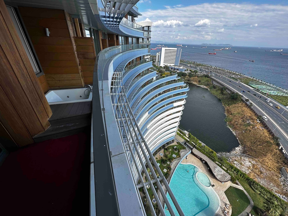 The multi-level balcony showcases a sweeping view of the sea and coastline. A private jacuzzi is positioned for relaxation, while a landscaped area with a pool is visible below. The architecture features curved lines and wooden accents, enhancing the visual appeal of the space.
