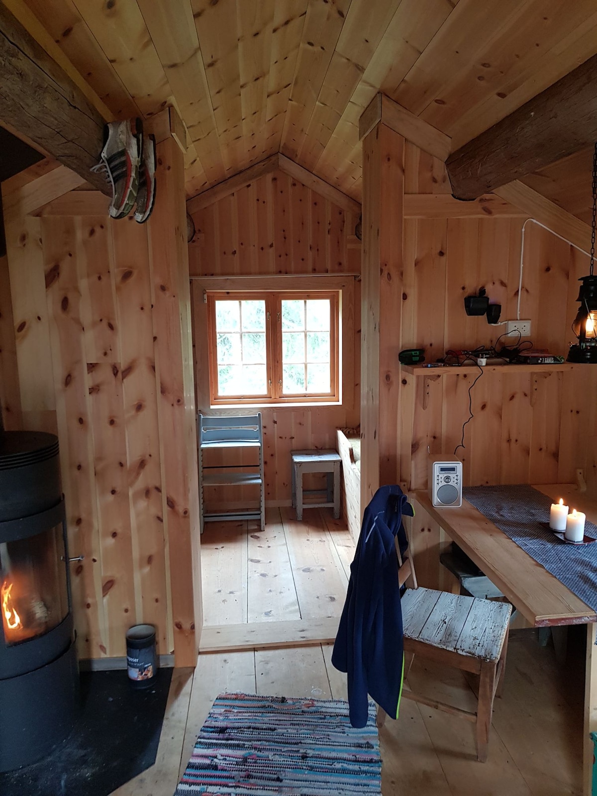 A wooden interior features natural timber walls and flooring, creating a warm ambiance. A small dining area with a wooden table and bench is visible. A lantern hangs beside the table, and a cozy stove is present, adding to the inviting atmosphere of the cabin.