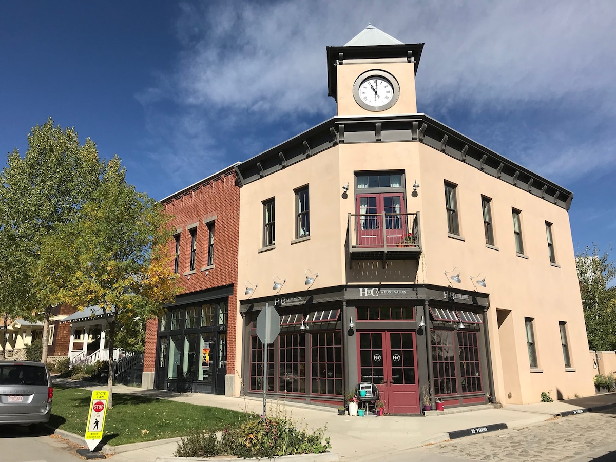 The Clocktower building, featuring a distinctive clock and a blend of brick and stucco architectural styles, stands prominently in the neighborhood. The ground floor has retail spaces with large windows, while the upper level offers a balcony with decorative planters under a clear blue sky.