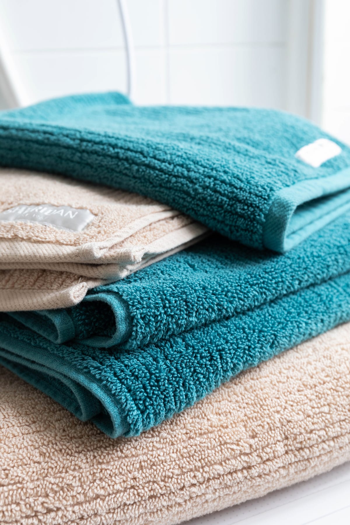A neatly arranged stack of soft towels showcases various textures and colors. The top towel is a deep teal, followed by a lighter beige towel, contributing to a cozy and clean aesthetic. The towels are placed on a white surface, with a hint of natural light illuminating them.