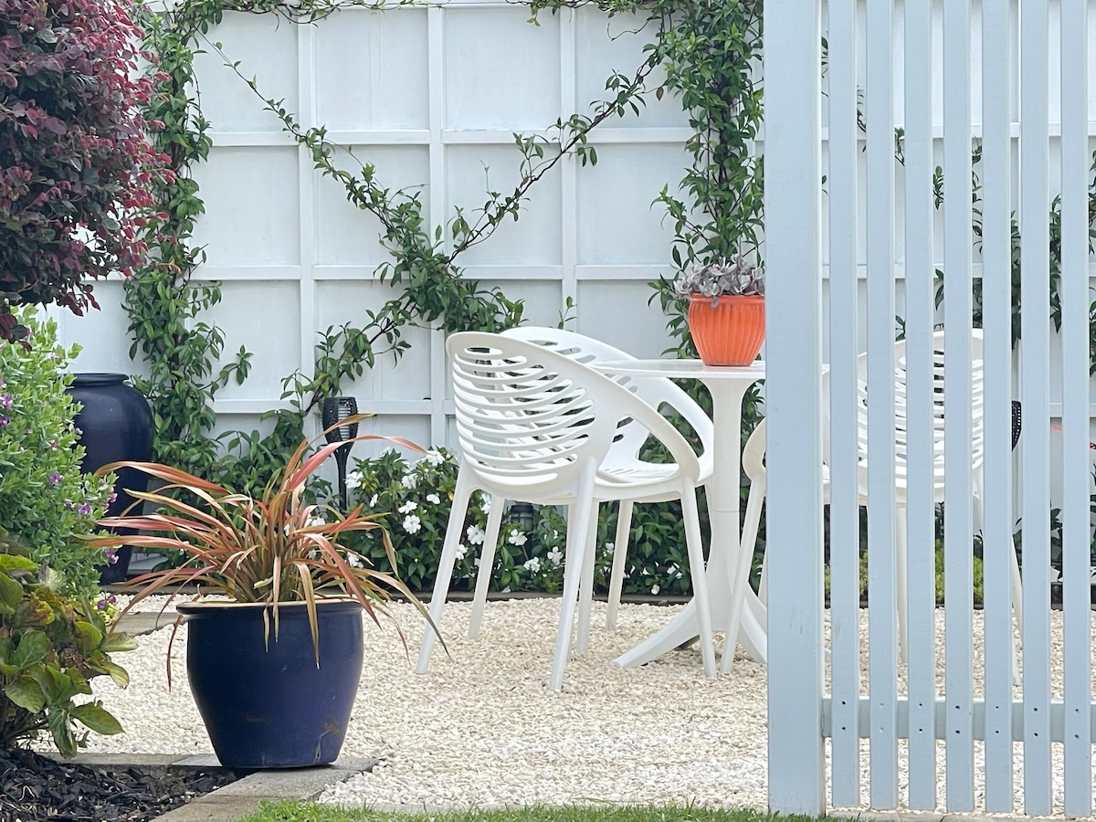 An outdoor seating area features a round white table surrounded by four white chairs. A vibrant orange pot holds a plant, contrasting with the lush green foliage on the trellis and the surrounding white gravel. The space offers a peaceful retreat amidst the garden.
