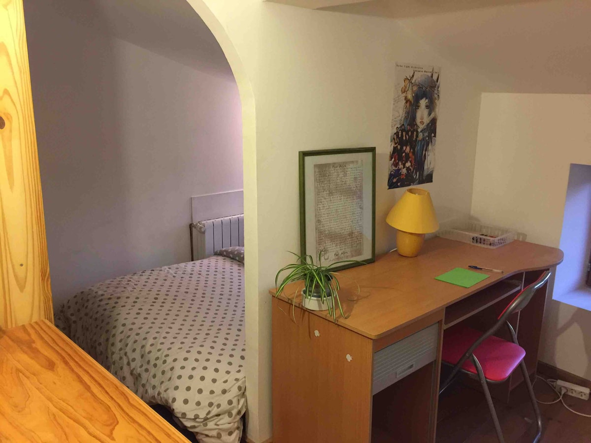 A compact room features a bed with a polka dot duvet cover set against a light-colored wall. A wooden desk with a bright lamp, a green plant, and a framed artwork occupies one side. A window allows natural light into the space.