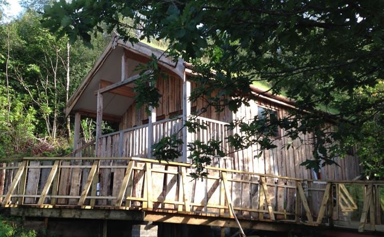 A unique wooden lodge is set on an elevated platform surrounded by greenery. The structure features a decked veranda with a railing, inviting outdoor access. Tall trees partially frame the view, enhancing the sense of seclusion in nature.