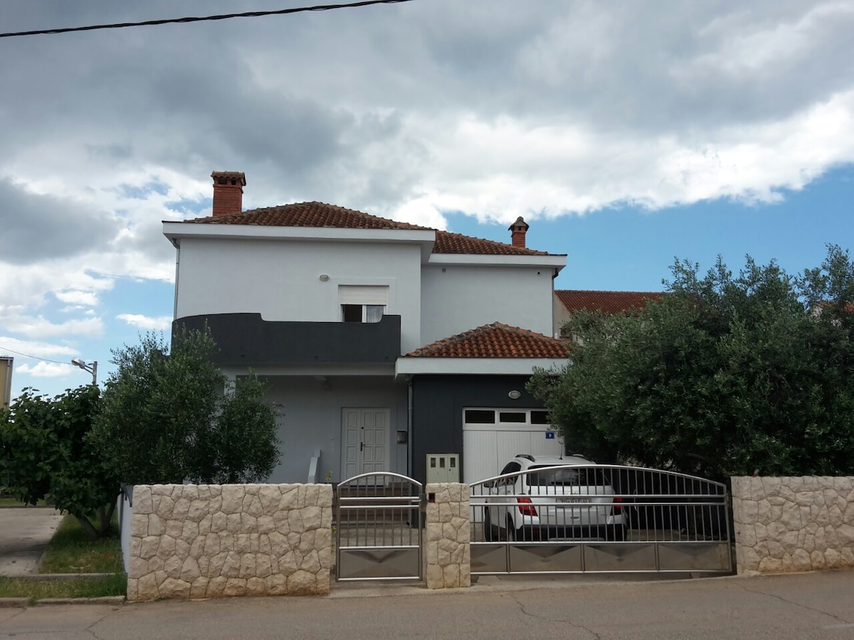 The exterior of a two-story house is presented, featuring a neutral color palette and a tiled roof. A gated entry leads to a driveway with a parked white car. Lush greenery surrounds the property, creating a welcoming environment. A cloudy sky adds a dramatic backdrop.