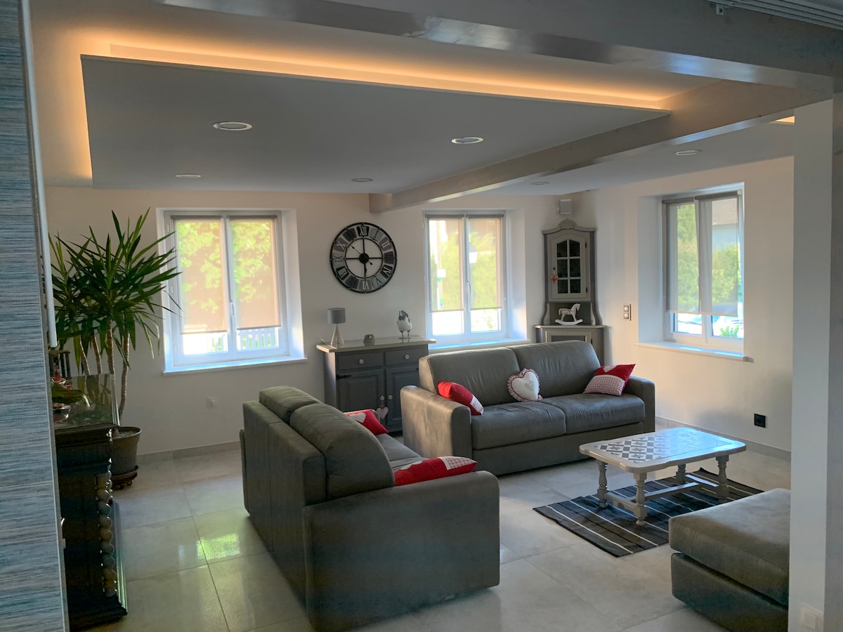 A spacious living area features neutral-toned sofas with red accent pillows. A modern clock is mounted on the wall above a dark sideboard. Large windows allow natural light to illuminate the room, enhancing the calming atmosphere. A patterned area rug lies atop the tile floor.