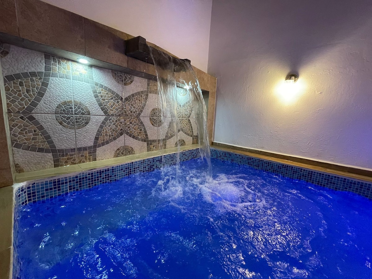 A serene pool area features a cascading waterfall that adds a soothing element to the environment. The pool's clear blue water shimmers under soft lighting, complemented by a decorative tiled wall that enhances the space's tranquil ambiance.