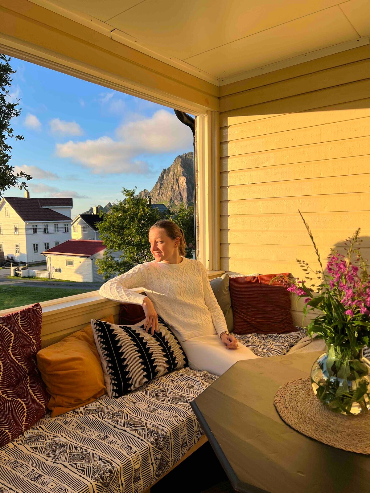 A cozy seating area is featured within a sunlit porch, adorned with colorful pillows and a patterned throw. A table, set with a vase of fresh flowers, invites relaxation while mountain views are visible through the large windows.