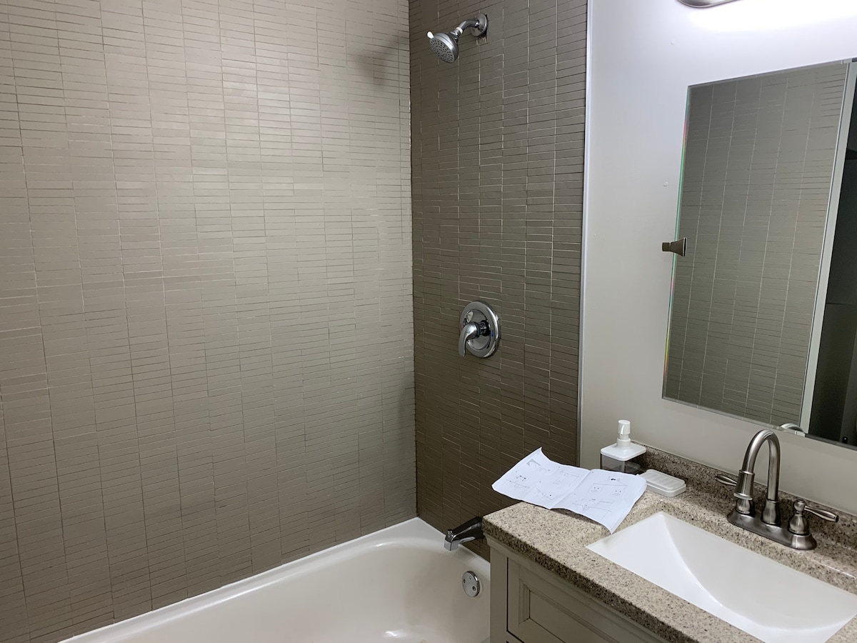 A modern bathroom features a spacious bathtub alongside a tiled shower enclosure. A neutral-toned wall is complemented by a granite countertop with a single sink and a large mirror above. Light fixtures provide bright illumination, enhancing the room's clean and contemporary design.