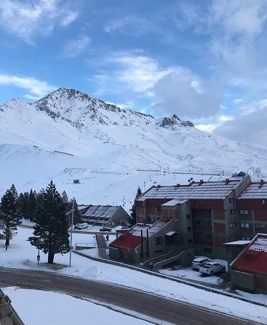 A snow-covered landscape showcases a mountain backdrop, with tiered buildings positioned in the foreground. Pine trees are visible, adding to the serene winter setting. The sky features a blend of blue and grey, suggesting a calm atmosphere over the snowy terrain.