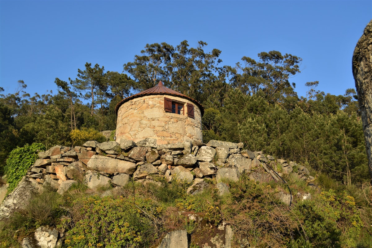 The historical windmill is perched on a rocky hillside, showcasing its traditional circular granite walls and distinctive conical roof. Surrounded by greenery, the building stands in harmony with its natural setting against a clear blue sky.