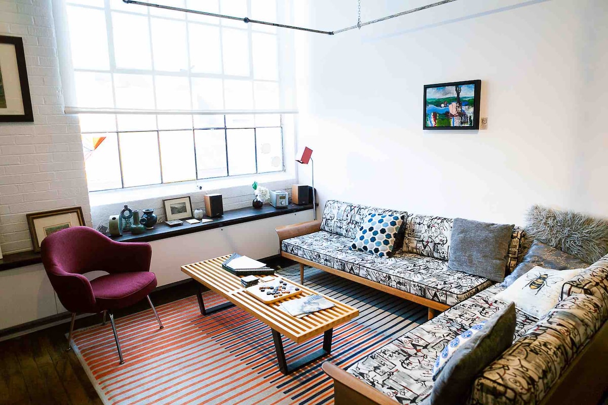 A cozy living area features a large window allowing natural light to fill the space. A colorful striped rug anchors the room, while a wooden coffee table sits in between two patterned sofas. A red accent chair adds a pop of color to the eclectic decor.