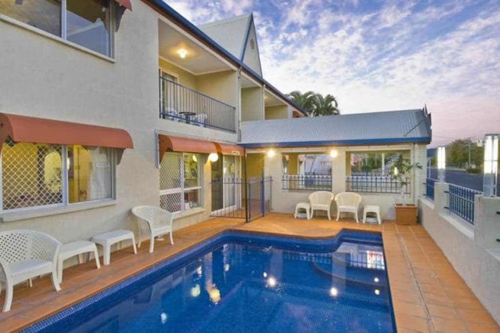 The outdoor area features a rectangular swimming pool with clear water, surrounded by beige tiled flooring. Four white lounge chairs are placed along one side of the pool, with two covered balconies visible above. A bright sky with scattered clouds adds to the inviting setting.