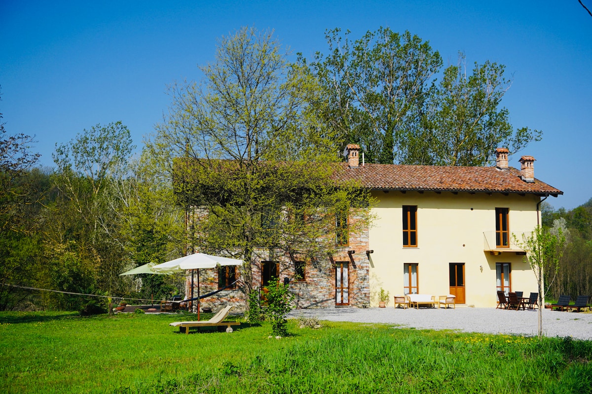 The exterior of the Casa al Tanaro is displayed, featuring a rustic two-story building with a warm color palette. Lush greenery surrounds the property, complemented by outdoor seating areas and umbrellas. A serene atmosphere is created by the spacious lawn and nearby trees.