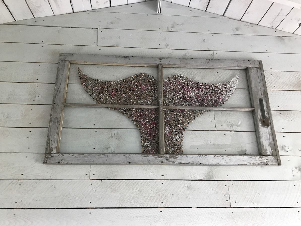 An artistic wall decoration is featured, composed of a vintage window frame filled with colorful textured materials arranged in the shape of a whale's tail. The frame is placed against a backdrop of whitewashed wooden panels.