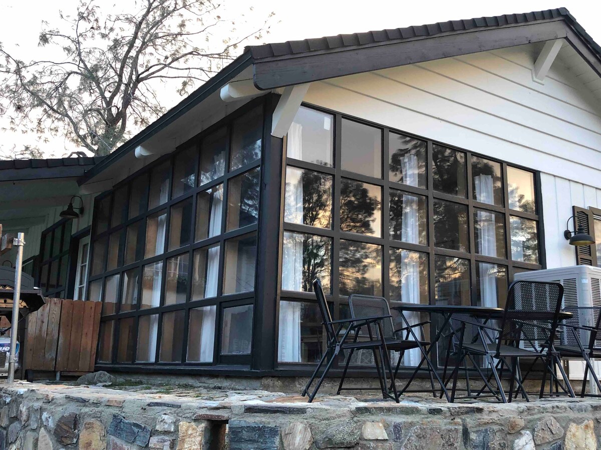 The exterior of the cottage features a sloped roof and a wall of large glass windows, providing a panoramic view of the surroundings. A stone patio area with a dining table and chairs is present, inviting outdoor relaxation.
