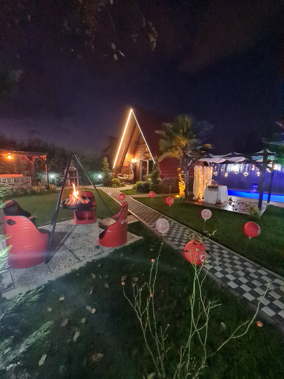 An inviting outdoor space is illuminated at night, featuring a cozy fire pit surrounded by red seating. A pathway lined with decorative stones leads to a wooden structure, while glowing lights enhance the ambiance. Lush greenery and palm trees create a relaxing atmosphere.