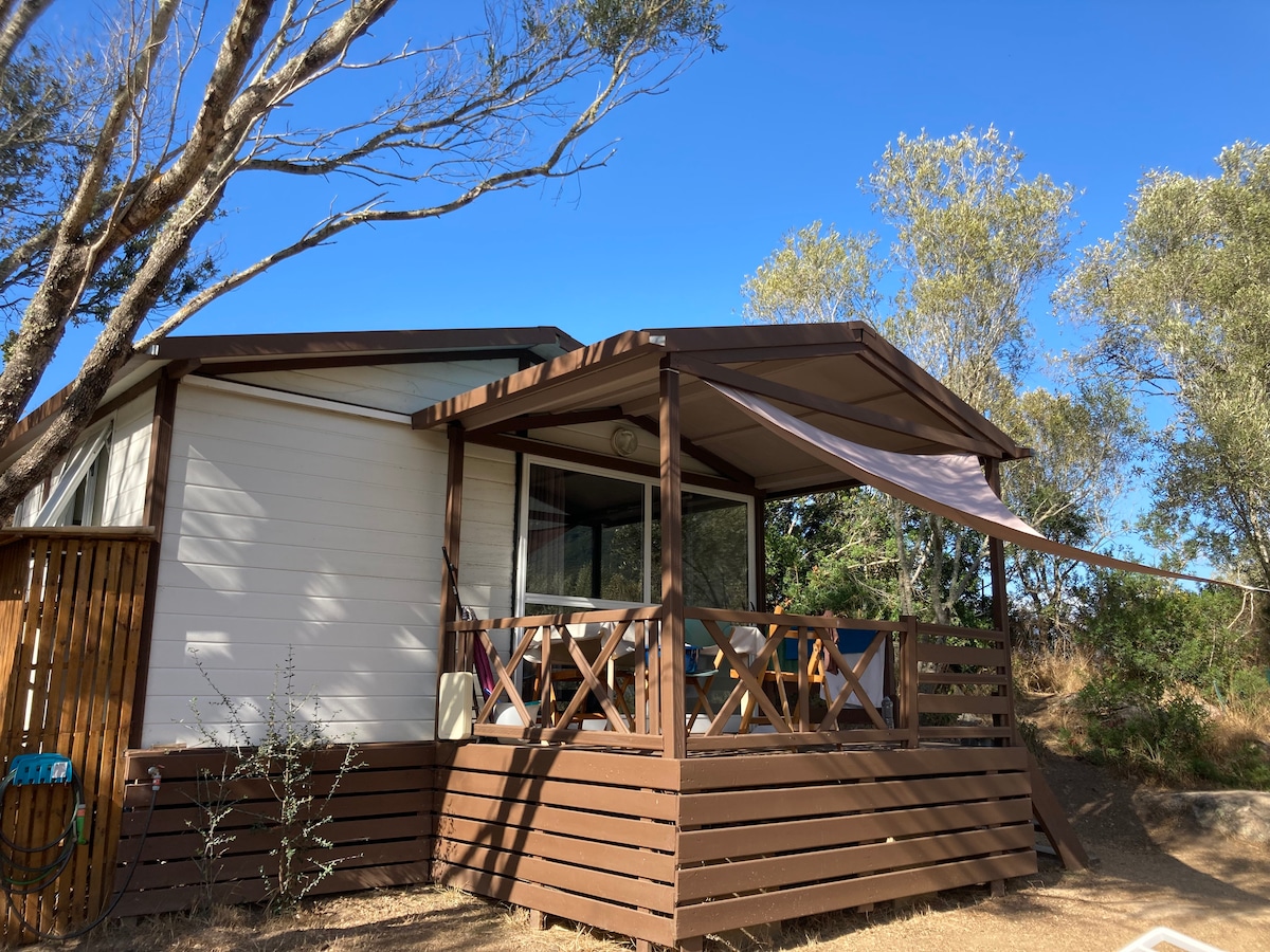 An inviting bungalow features a covered wooden porch with a railing, offering outdoor seating. Large windows allow natural light to enter the interior. Surrounding trees provide a serene atmosphere, while the clear blue sky complements the peaceful outdoor setting.