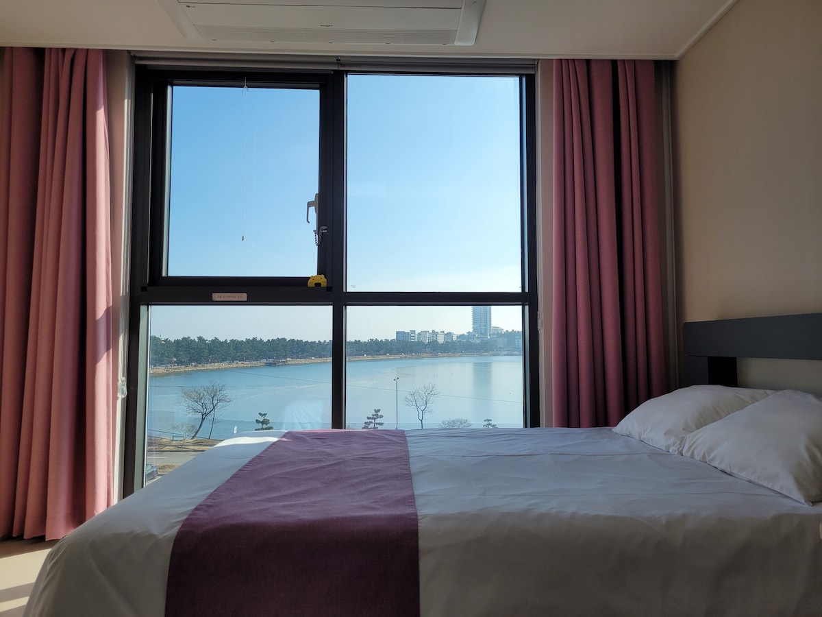A spacious bedroom features a large window with views overlooking a serene body of water. Soft pink curtains frame the window, allowing natural light to fill the room. A neatly made bed with a lavender bedspread rests against the wall, inviting relaxation.