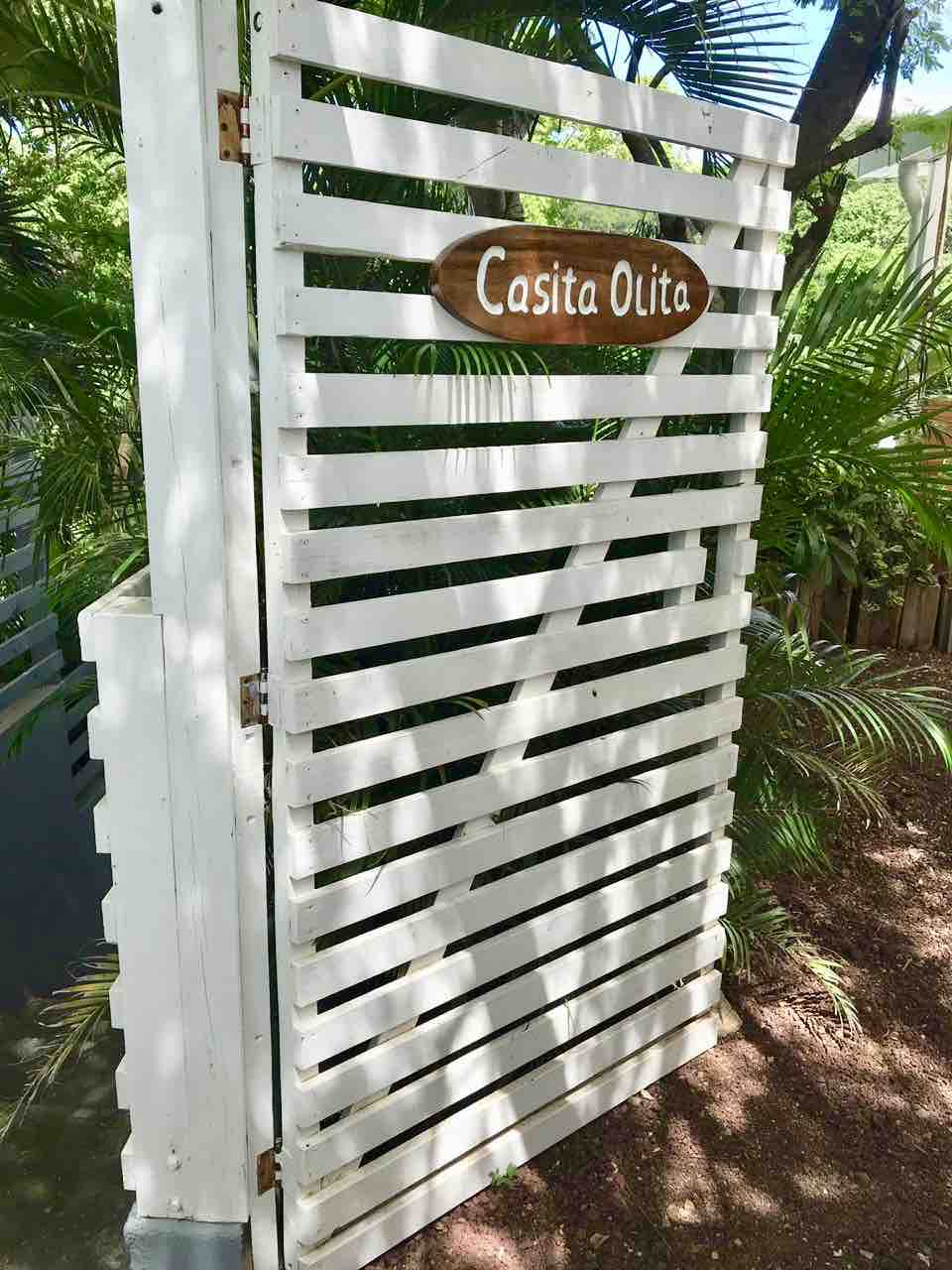 A white wooden gate with horizontal slats is visible, featuring a carved wooden sign that reads 'Casita Olita.' Lush green plants surround the gate, contributing to a tropical setting.