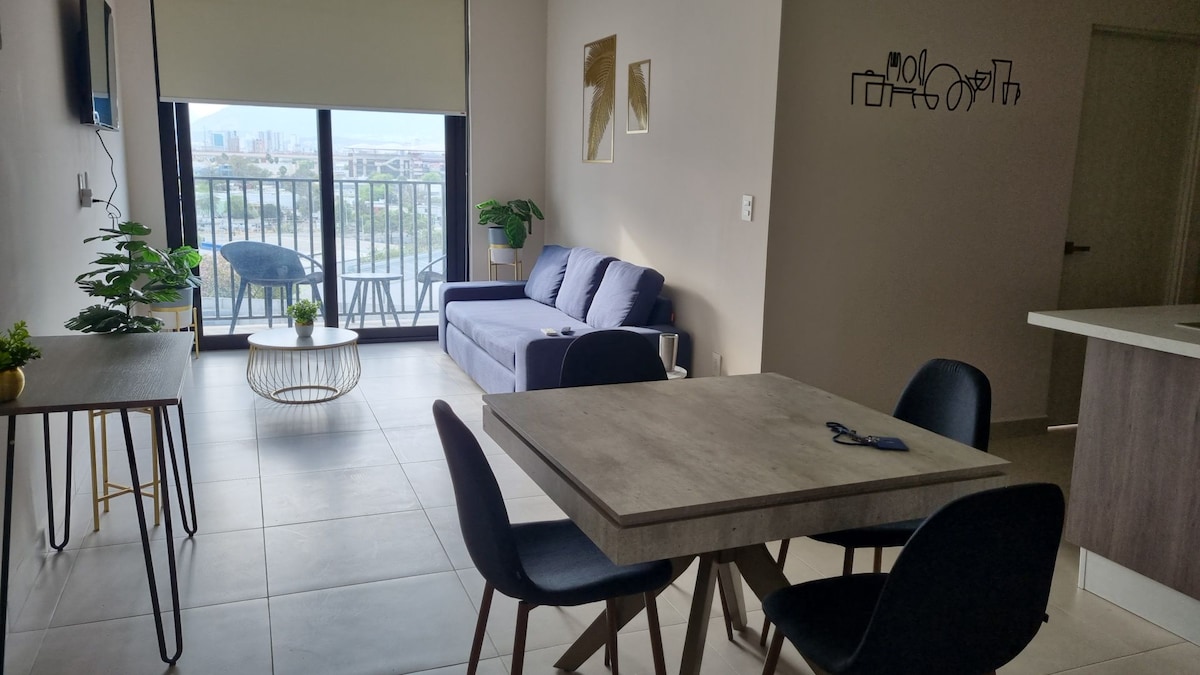 A modern living area is visible, featuring a blue sofa and a dining table set with four chairs. Natural light floods the space through large glass doors opening to a balcony, while potted plants add a touch of greenery.