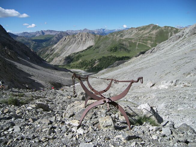 The rugged landscape features a rocky terrain with steep mountain slopes and green patches in the distance. A rusted metal structure rests on the ground, surrounded by scattered boulders and sparse vegetation. A clear blue sky with a few clouds enhances the expansive view.