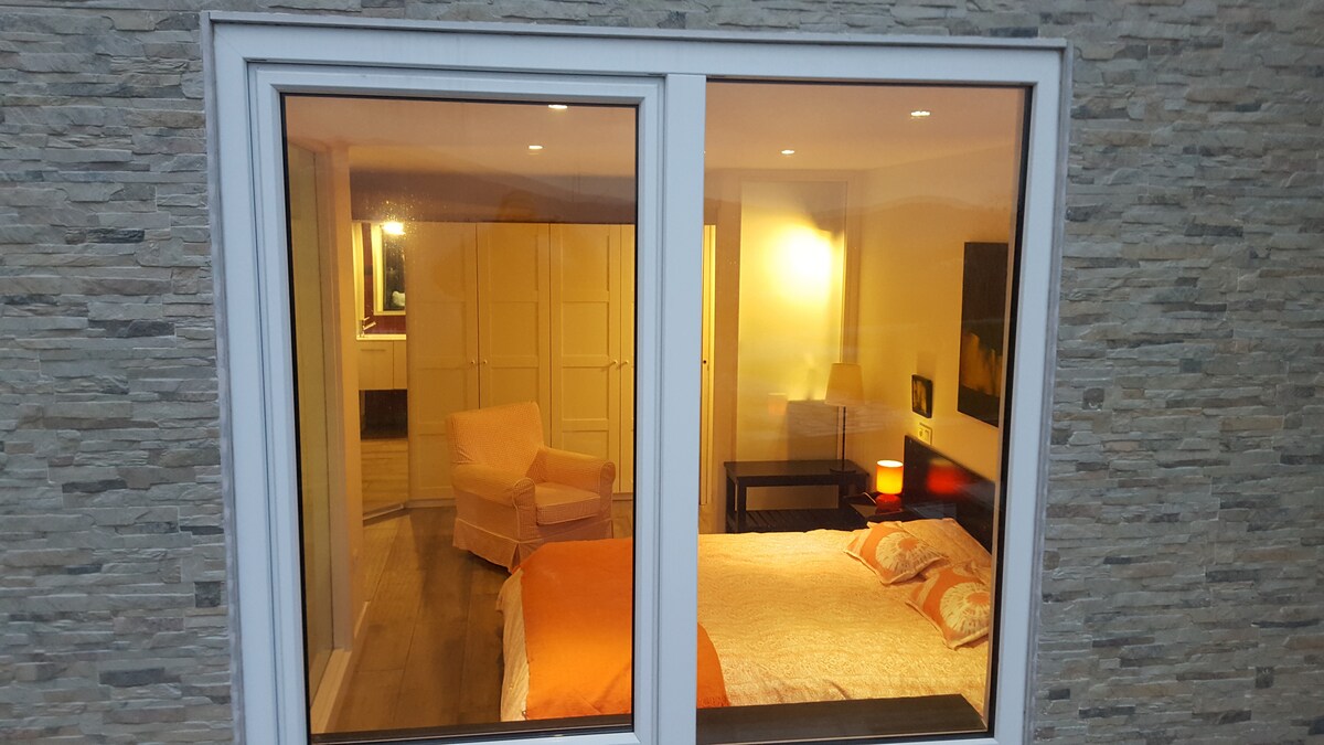 A cozy bedroom reveals a spacious bed adorned with orange accents and decorative pillows. A chair sits in one corner, while soft lighting creates a warm atmosphere. Sliding glass doors provide a view into the room’s inviting design.
