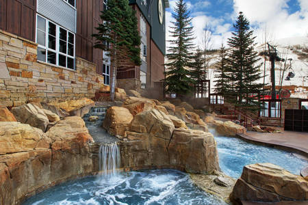 The outdoor area features a rocky landscape with a hot tub surrounded by natural stone. Water cascades gently from the rocks, creating a tranquil atmosphere. Lush evergreen trees frame the scene, with the ski resort's exterior visible in the background against a partly cloudy sky.