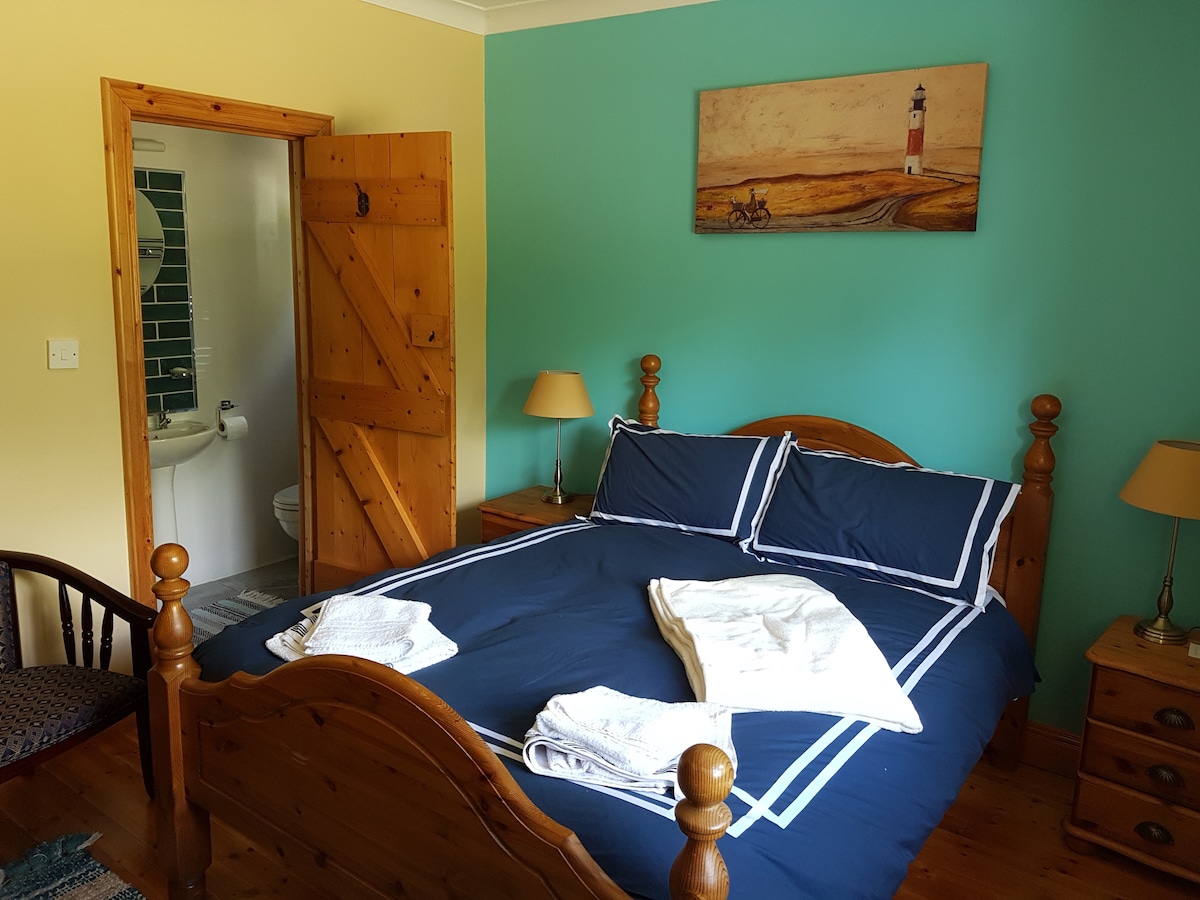 A bright bedroom features a wooden bed with navy blue bedding neatly arranged. Light-colored bedside tables with lamps stand beside the bed. A painting of a lighthouse adorns the wall above the bed. An ensuite bathroom is visible through an open door.