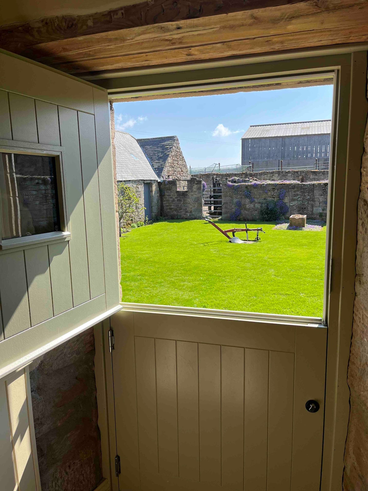 A partially open door reveals a well-maintained outdoor area with lush green grass. Stone walls and rustic structures define the space, while a bench and other features are visible in the distance under a clear blue sky.