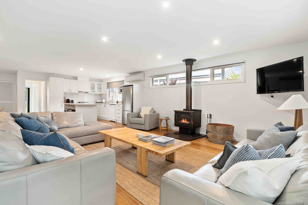 An open plan living area features light-colored sofas arranged around a wooden coffee table. A cozy log fireplace is situated against a white wall, with a television mounted nearby. Bright light filters in through large windows, enhancing the modern aesthetic.