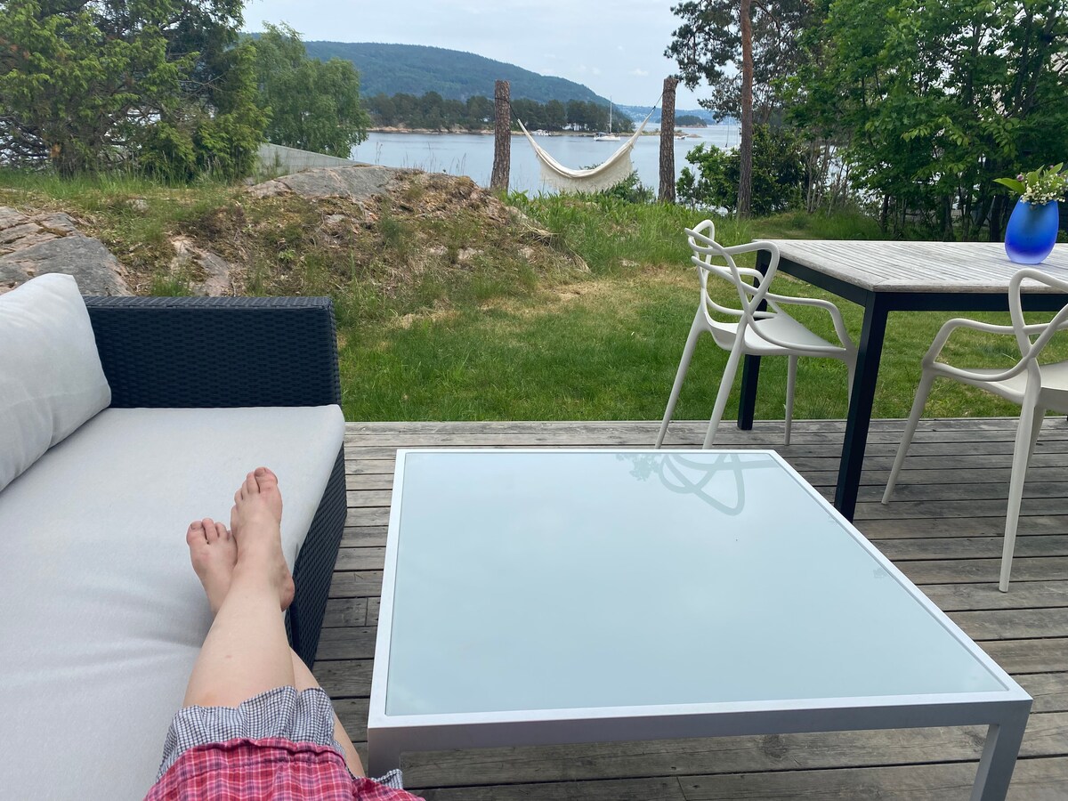 A comfortable outdoor seating area features a sectional sofa and a glass-top table. In the background, a hammock is seen hanging between trees, and a calm water view is visible, surrounded by greenery and distant hills.