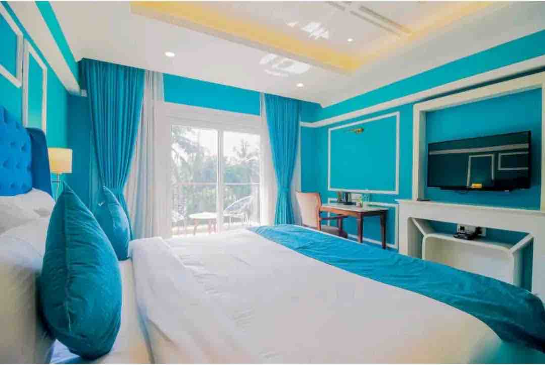 A bright sleeping area features vibrant blue walls, complemented by light curtains and decorative pillows. A spacious bed with crisp white linens is positioned in front of a large glass door that opens to a balcony. A television is mounted on the wall opposite.