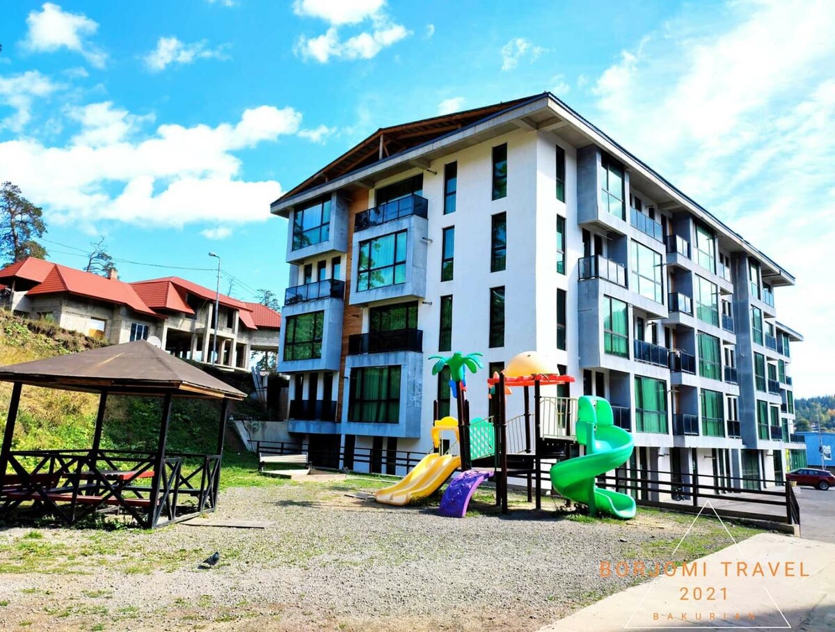 The apartment building features a modern design with multiple large windows that allow for ample natural light. An outdoor play area with a playground, slides, and climbing structures is visible, offering a space for children to play. A shaded seating area with wooden benches is also present.