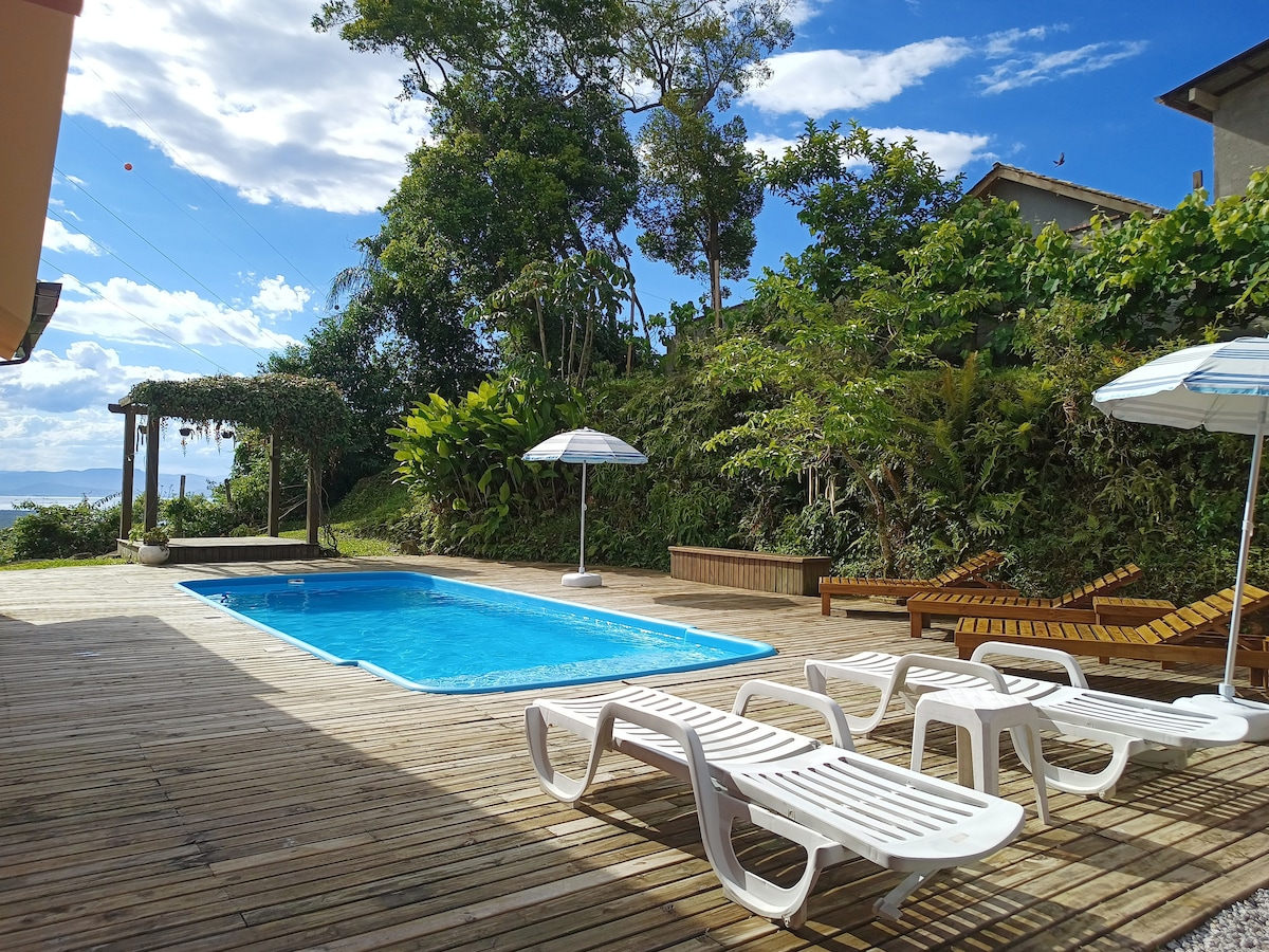 A wooden deck surrounds a swimming pool that reflects the blue sky. Lounge chairs are placed nearby, under umbrellas, while lush greenery creates a natural backdrop, suggesting a serene outdoor space for relaxation and enjoyment.