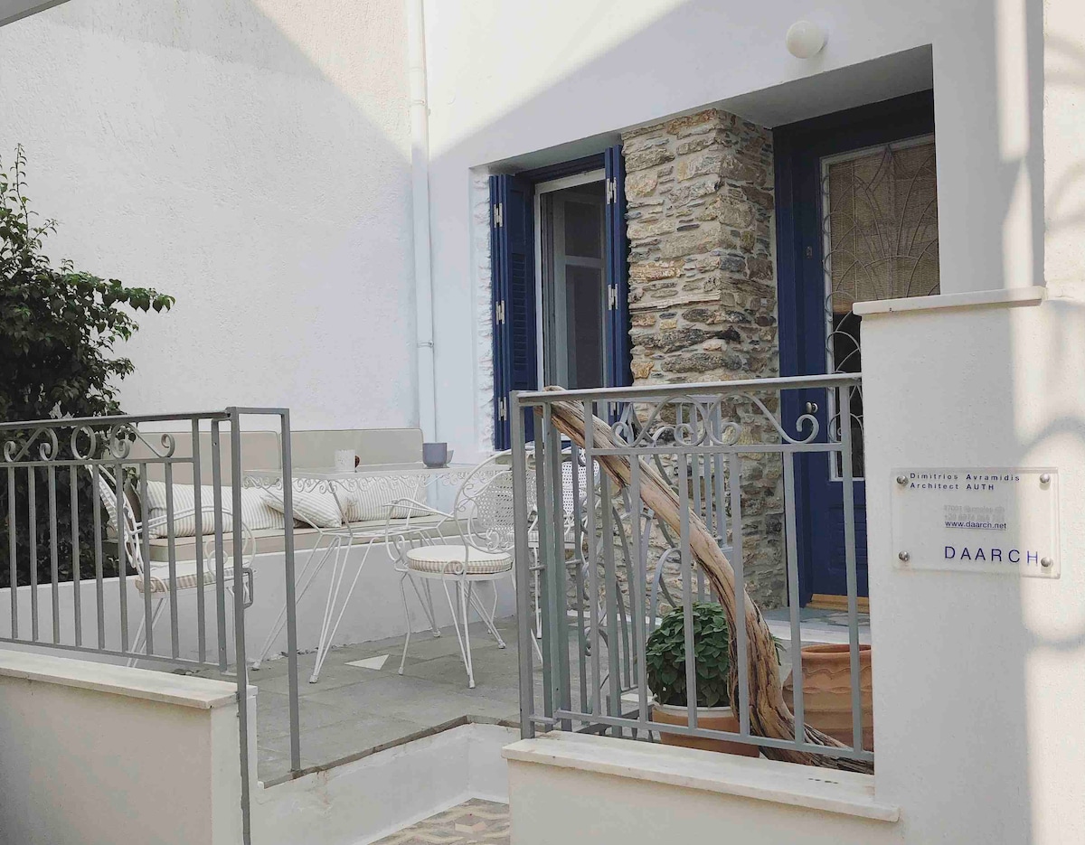 A charming entrance is framed by a stone wall and dark blue shutters, leading to a private seating area. White wrought-iron furniture is arranged on a tiled patio, surrounded by greenery, contributing to a welcoming outdoor space.