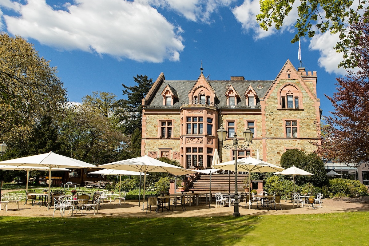 A historic building with ornate architectural details is surrounded by green trees and well-maintained gardens. Outdoor seating areas feature white umbrellas over tables, inviting guests to relax in the serene environment. A staircase leads to the entrance, framed by lush floral arrangements.