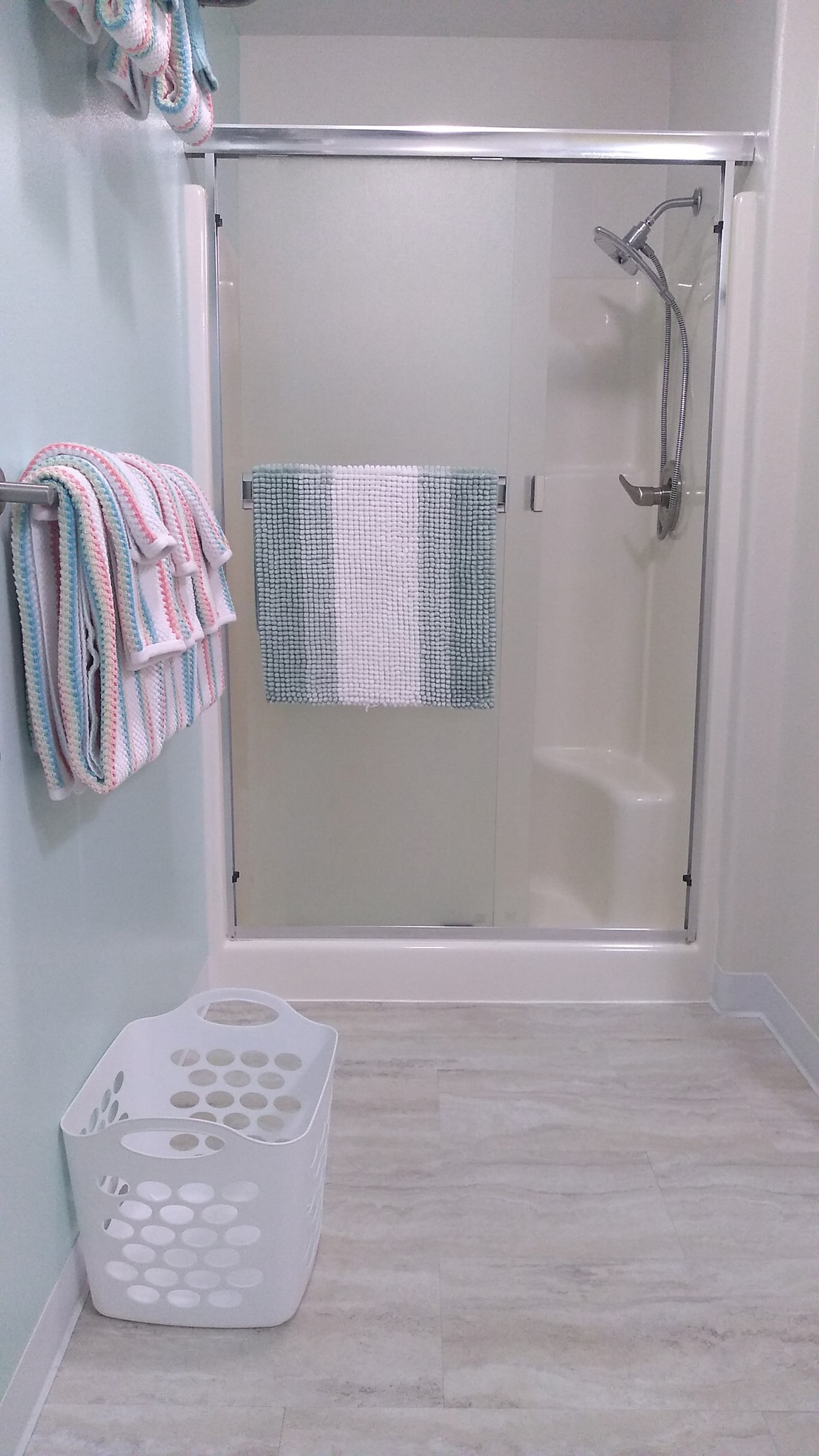 A modern bathroom features a step-in shower with a glass door. Soft towels are neatly hung on a bar, while a striped towel decorates the shower space. A laundry basket is positioned on the floor, complementing the light-colored tiles and pastel walls.