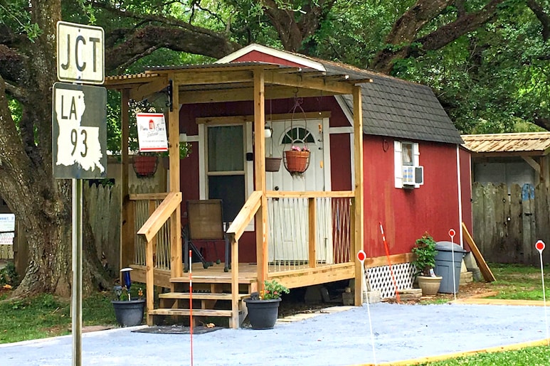 A converted red barn shed features a covered front porch with a wooden rail. Potted plants adorn the porch, enhancing the rustic exterior. The structure is surrounded by greenery, including large oak trees, contributing to a serene atmosphere.