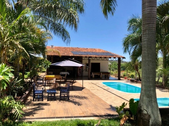 A sunny outdoor area features a swimming pool beside a shaded patio. A dining table with chairs is placed under an umbrella, while lounge chairs line the side. Lush palm trees surround the space, enhancing the tropical atmosphere of the backyard.