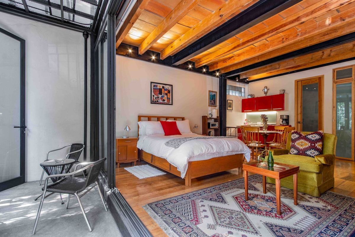A spacious studio features a comfortable bed with white linens and a red accent pillow. Warm wooden beams frame the ceiling, while a cozy seating area includes a green chair and patterned rug. Large glass doors open to a private terrace, offering natural light and inviting views.