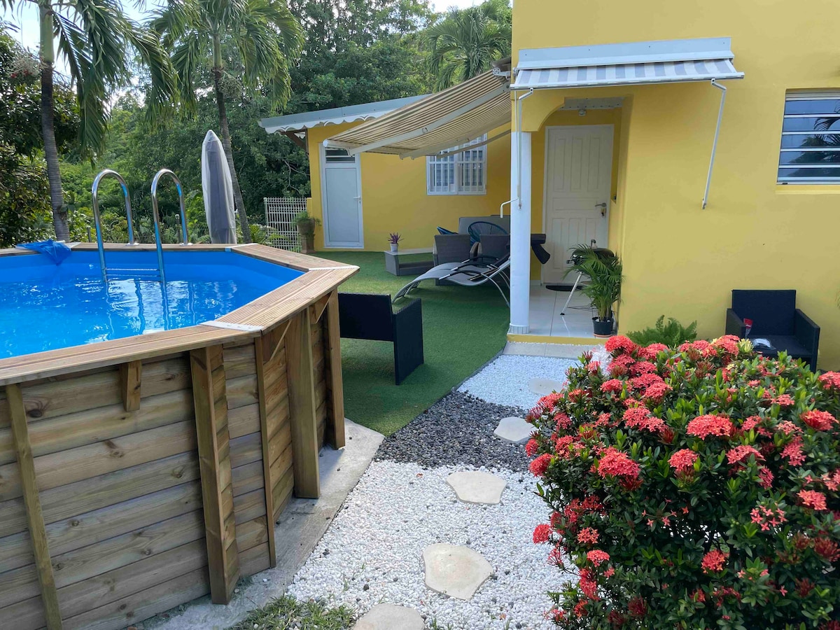 A spacious outdoor area features a round blue pool surrounded by a wooden deck, complemented by green grass. A yellow building provides shade, while lounge chairs and a small table are positioned nearby. Colorful flowers and stepping stones add to the serene garden ambiance.