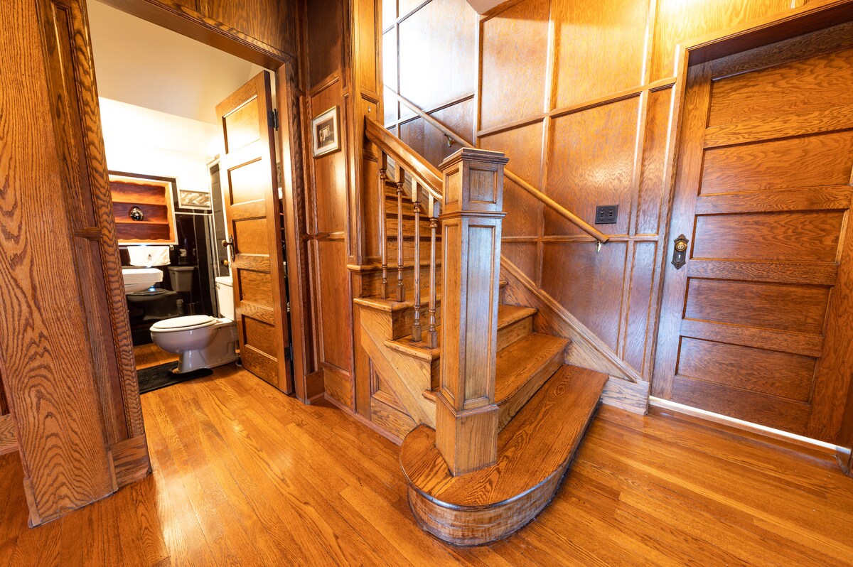 A wooden staircase with a curved design leads to the upper level, surrounded by rich wood paneling. The warm, polished floor adds a cozy touch, while several doors provide access to different areas, including a glimpse of a bathroom. Natural light brightens the inviting entrance.