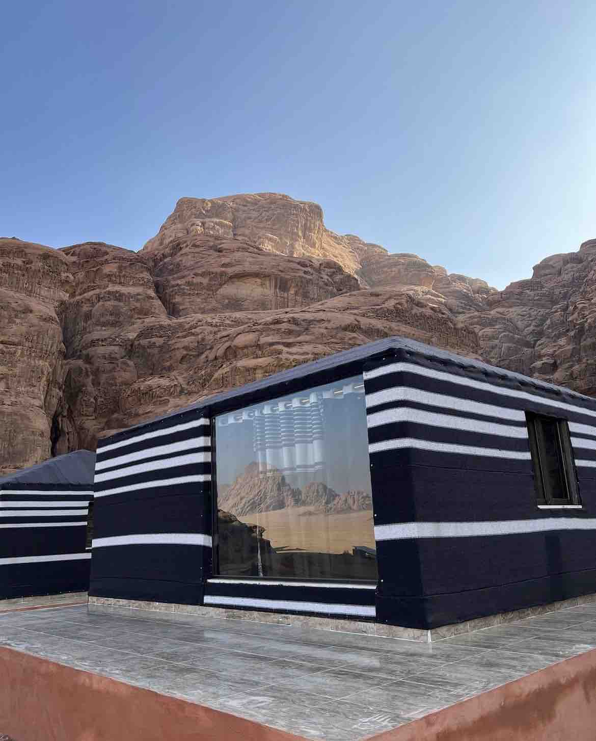 A distinctive black and white striped cabin is positioned against a backdrop of towering rock formations. Large windows allow natural light to flood the interior, showcasing the beauty of the surrounding landscape. The deck area features wooden flooring, complementing the cabin's unique design.
