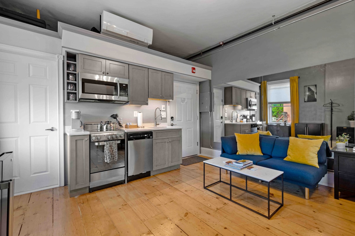 A modern studio kitchen features stainless steel appliances, including a microwave and dishwasher, set against grey cabinetry. A light wood floor adds warmth to the space, complemented by a blue sofa and a simple coffee table. Natural light enters from a nearby window, enhancing the bright atmosphere.
