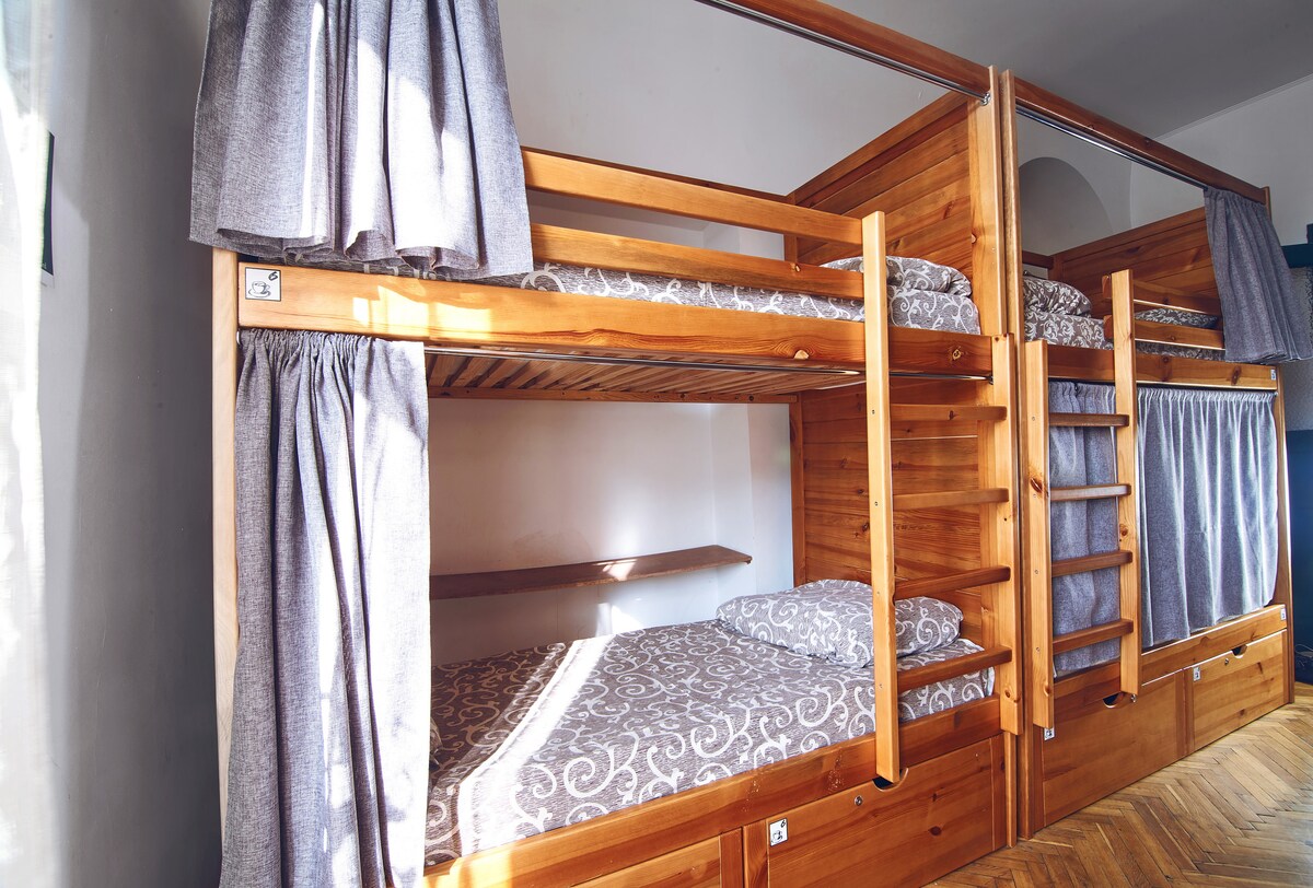 A dormitory room features wooden bunk beds with curtains for privacy. Each bunk has a patterned mattress, and a small shelf is located between the beds. Light filters in through a nearby window, creating a bright and inviting atmosphere.