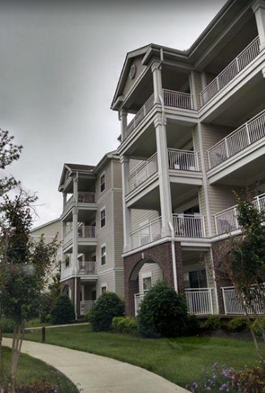 A multi-story building exhibits a mix of light-colored siding and brick accents. Several balconies extend from the upper levels, framed by white railings. Well-manicured green grass and small shrubs line the pathway leading to the entrance.