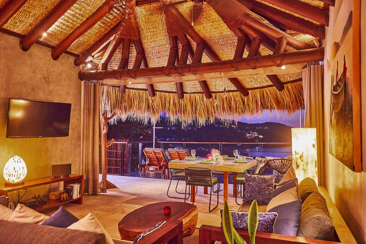 A spacious living area is featured under a thatched roof, showcasing a blend of natural materials. Sliding glass doors open to a scenic outdoor dining space. Soft lighting and comfortable furnishings create an inviting atmosphere, while a television is mounted on the wall.
